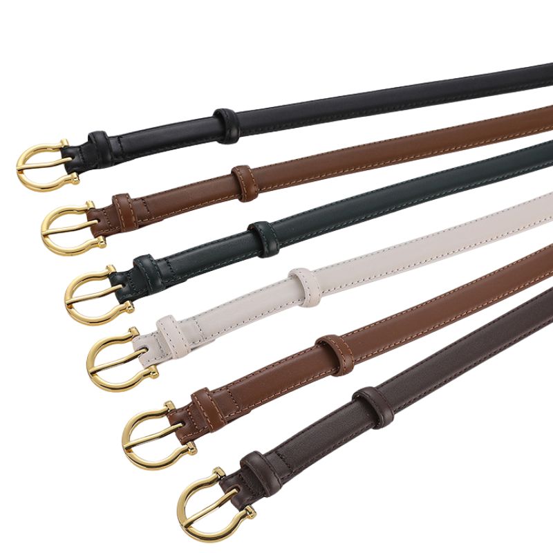 Six women's Cassandre 1 belts in black, brown, dark green, white, medium brown, and dark brown, each with a slim leather strap and a gold half-moon buckle.